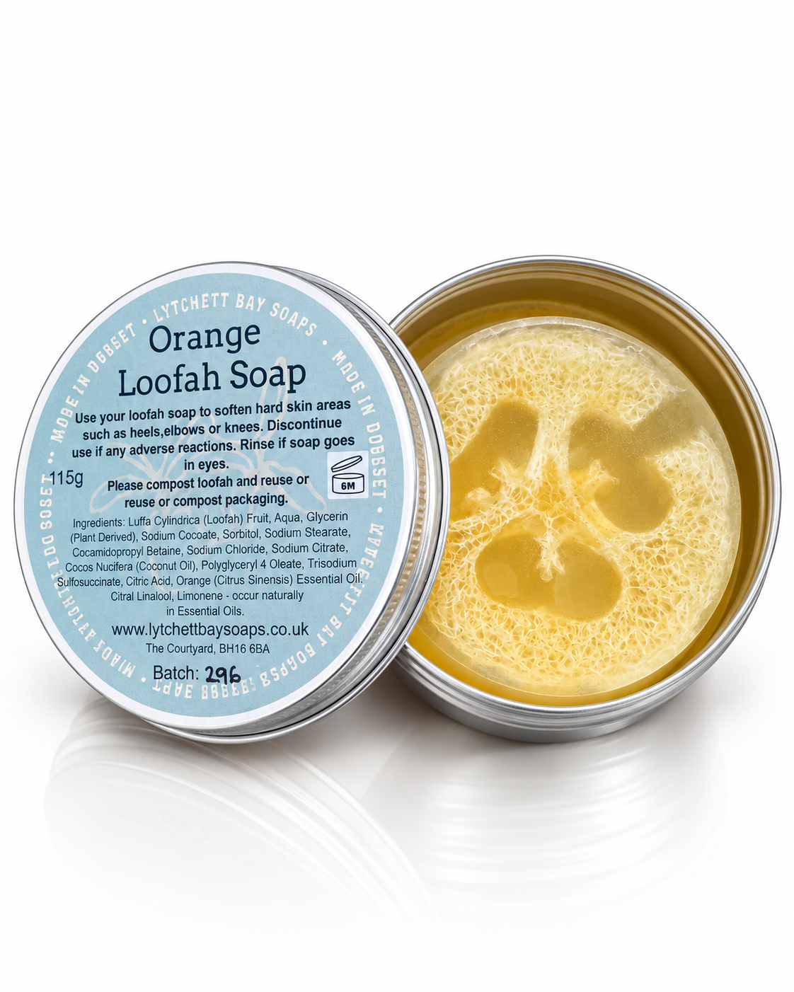 An open round metal tin of Lytchett Bay Soaps Orange Loofah Soap, showing the pale yellow loofah inside, with the lid placed beside it on a white reflective surface.